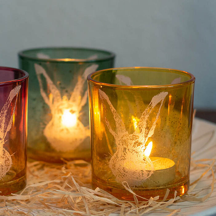 Drei brennende Duftkerzen des Herstellers "Wurm" in bunten Glasbehältern (rosa, grün und orange). Die Kerzen sind auf einem weißen Keramikteller platziert, der mit Stroh ausgelegt ist. Im Hintergrund steht eine rosa Vase mit getrockneten Blumen. Die Glasb