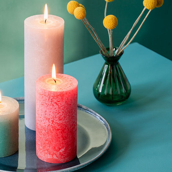 Sechs brennende Bolsius Rustikkerzen in verschiedenen Größen und Farben, darunter Weiß, Grün und Rosa, stehen auf einer Holzablage. Daneben befinden sich eine rosa Blume in einer kleinen Vase und ein Set aus Keramikgeschirr. Die Szene wirkt warm und einla
