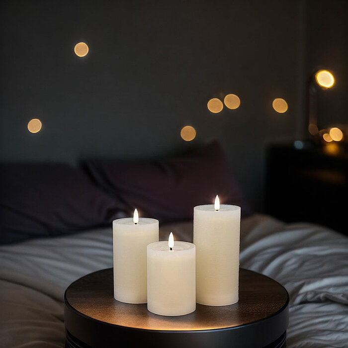 Drei brennende, weiße Stumpenkerzen stehen auf einem runden, dunklen Holztisch. Die Szenerie ist in ein warmes, gemütliches Licht getaucht. Im Hintergrund sind ein Bett mit dunklen Kissen und eine leicht beleuchtete Nachttischlampe zu sehen. An der Wand s