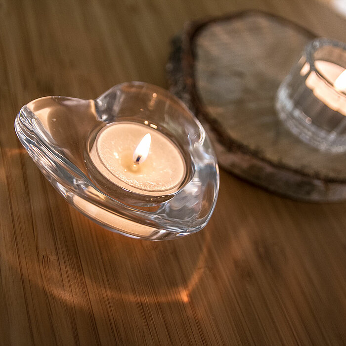 Ein brennendes Teelicht in einem herzförmigen Teelichthalter und ein weiteres brennendes Teelicht in einem zylindrischen, klaren Glasbehälter auf einer runden Holzscheibe stehen auf einer Holzoberfläche. Die Produkte sind vom Hersteller "Sandra Rich".