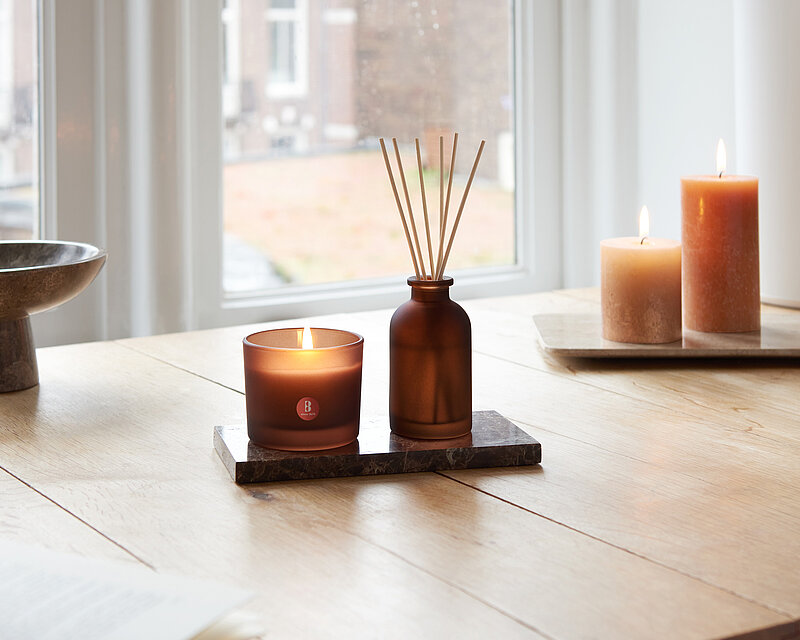 Ein heller, gemütlicher Wohnraum mit einem Holztisch. Auf dem Tisch stehen ein Bolsius Duftglas True Joy und ein Raumduft-Diffuser mit Rattanstäbchen aus braunem Milchglas.