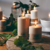 Neun brennende Kerzen des Herstellers Wenzel in verschiedenen Größen. Die Kerzen bestehen aus natürlichem Wachs und tragen das Label "PURE NATURAL WAX". Sie stehen auf einem Holz- und einem Steintablett, welches mit grünen Blättern dekoriert ist. Im Hinte