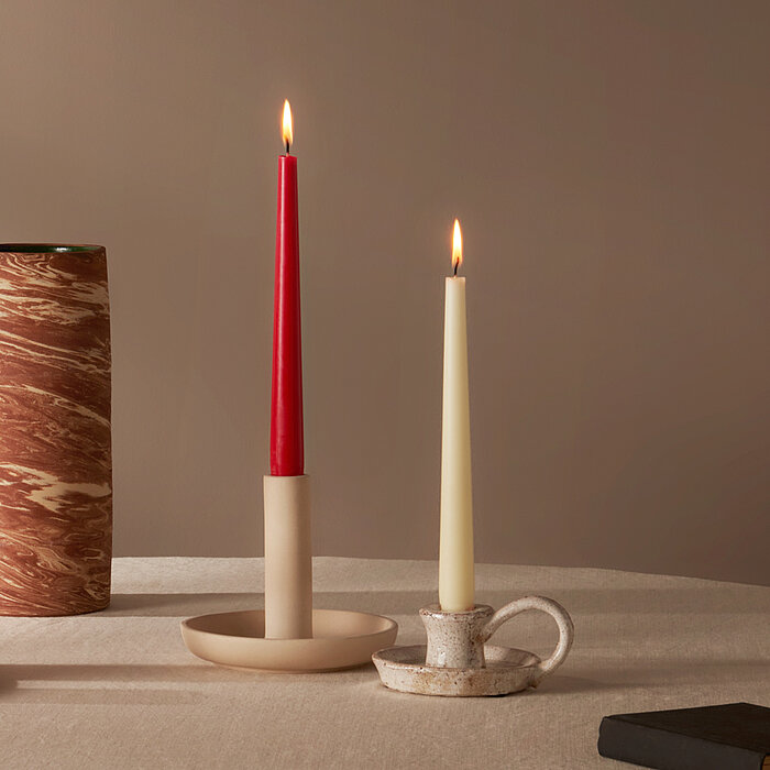 Zwei brennende Bolsius Tafelkerzen, eine rote und eine elfenbeinfarbene, in unterschiedlichen Kerzenhaltern auf einem runden Tisch. Auf dem Tisch befinden sich zudem eine beige und eine rote Stumpenkerze von Bolsius auf einem runden Tablett, sowie ein sch