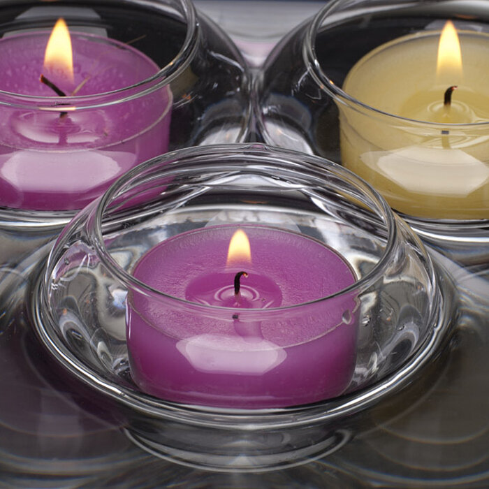 Vier brennende Teelichter vom Hersteller Sandra Rich. Zwei in violetten Kerzen in runden, durchsichtigen Glasbehältern, eine gelbe Kerze in einem ähnlichen Glasbehälter. Sie stehen auf einer Oberfläche, die das Licht reflektiert und eine beruhigende Atmos