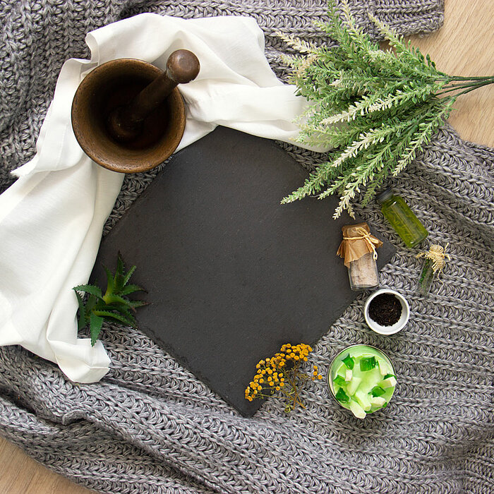 Eine graue Strickdecke auf einem hellen Holzboden bildet den Hintergrund. Darauf liegt ein dunkles, quadratisches Schieferbrett. Rechts oben sind grüne Kunstpflanzen zu sehen. Links oben befindet sich ein Mörser mit Stößel aus Holz, daneben ein weißes Tuc