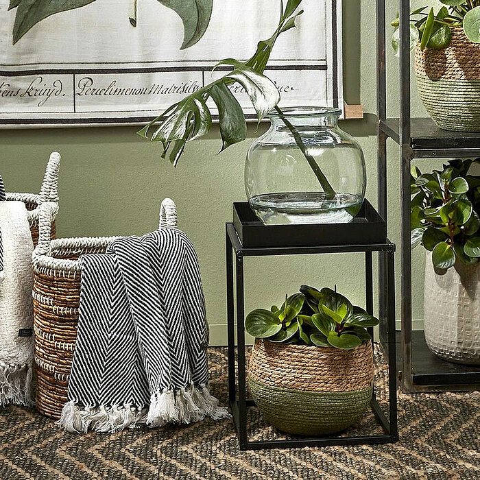 Drei geflochtene Körbe, zwei in einem gestreiften Muster und einer in grün, gefüllt mit zwei grau-weiß gestreiften Decken, stehen auf einem gemusterten Teppich. Neben den Körben befindet sich ein schwarzer Beistelltisch mit einer großen Glasvase, aus der 