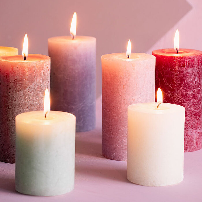 Drei brennende Bolsius Rustikkerzen in verschiedenen Größen und Farben, die auf einem runden weißen Tablett stehen. Die Farben der Kerzen sind ein tiefes Rot, ein mittleres Rosa und ein blasses Rosa. Im Hintergrund ist ein gemütliches Wohnzimmerambiente m