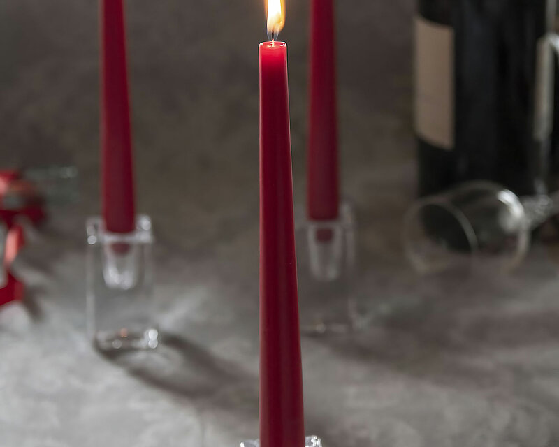 Auf einer grauen mit weißen Ornamenten verzierten Tischdecke stehen drei eckige Glas-Stabkerzenhalter mit je einer alt roten brennenden Spitzkerze von Bolsius. Im Hintergrund erkennt man am rechten Bildrand eine Flasche Wein und ein liegendes Weinglas. Am rechten Bildrand im Hintergrund befinden sich einige rote Blüten.