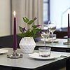 Zwei brennende lilafarbene Stabkerzen stehen in silbernen Kerzenhaltern auf einem gedeckten Esstisch. Auf dem Tisch befinden sich zudem Weingläser, Wassergläser, weiße Servietten und eine weiße Vase mit lila Blumen. Im Hintergrund sind weitere gedeckte Ti