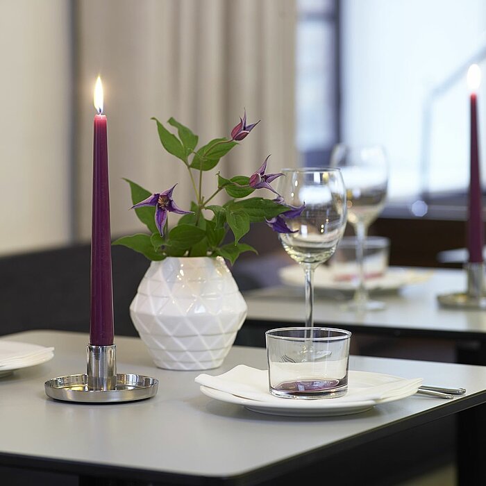 Zwei brennende lilafarbene Stabkerzen stehen in silbernen Kerzenhaltern auf einem gedeckten Esstisch. Auf dem Tisch befinden sich zudem Weingläser, Wassergläser, weiße Servietten und eine weiße Vase mit lila Blumen. Im Hintergrund sind weitere gedeckte Ti