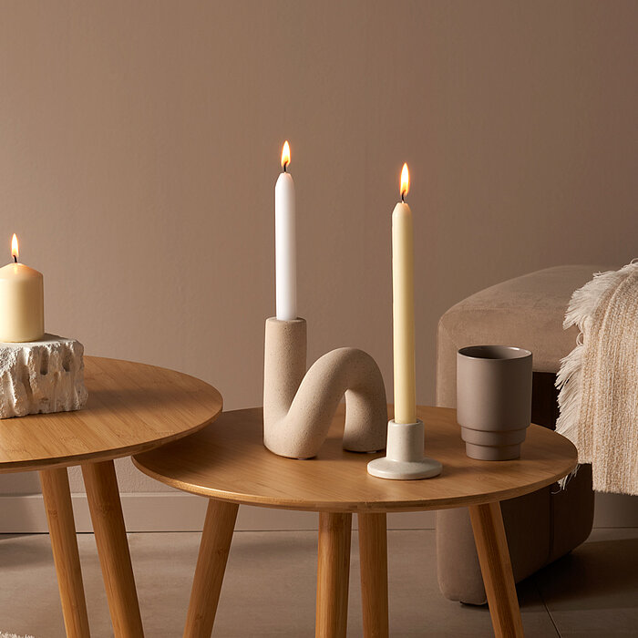 Vier brennende Bolsius Haushaltskerzen in verschiedenen Formen und Größen. Auf einem runden Holztisch stehen zwei Kerzenhalter mit einer weißen und einer gelben langen Kerze, daneben steht eine beige Stumpenkerze auf einem Steinsockel. Auf einem kleineren
