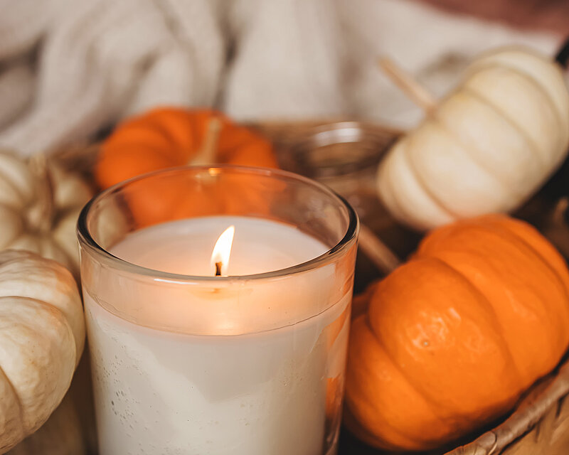 Nahaufnahme eines brennenden Duftkerzenglases in einem geflochtenen Korb-Tablett. Das Glas ist umgeben von weißen und orangenen Kürbissen.