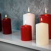 Sechs brennende DecoLite Kerzen in den Farben Rot und Weiß stehen in verschiedenen Größen auf einem weißen Regal vor einer grauen, gemusterten Tapete. Die Stimmung ist ruhig und festlich.