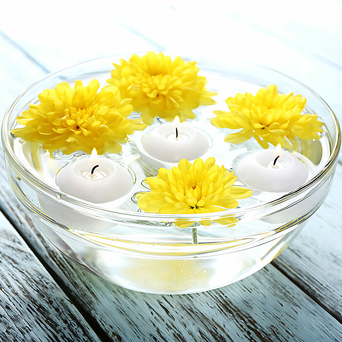 Eine Glasschüssel mit Wasser, in der drei weiße schwimmende Kerzen und vier gelbe Chrysanthemenblüten schwimmen. Die Produkte von DecoLite erzeugen eine ruhige und friedliche Atmosphäre auf einem weißen, verwitterten Holztisch.