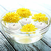 Eine Glasschüssel mit Wasser, in der drei weiße schwimmende Kerzen und vier gelbe Chrysanthemenblüten schwimmen. Die Produkte von DecoLite erzeugen eine ruhige und friedliche Atmosphäre auf einem weißen, verwitterten Holztisch.