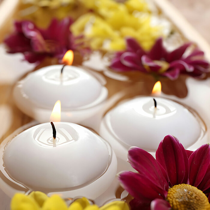Drei schwimmende weiße Kugelkerzen von DecoLite brennen auf einer Wasseroberfläche, umgeben von roten und gelben Blütenblättern. Die Szene vermittelt eine ruhige und entspannende Atmosphäre.
