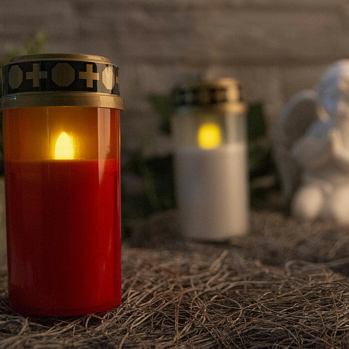 Ein Bild zeigt eine elektrische Kerze von ARTS in einem roten Gehäuse mit einem goldenen Deckel, der mit Kreuzen und Kreisen verziert ist. Im Hintergrund steht eine weitere elektrische Kerze von ARTS in einem weißen Gehäuse. Beide Kerzen stehen auf einer 