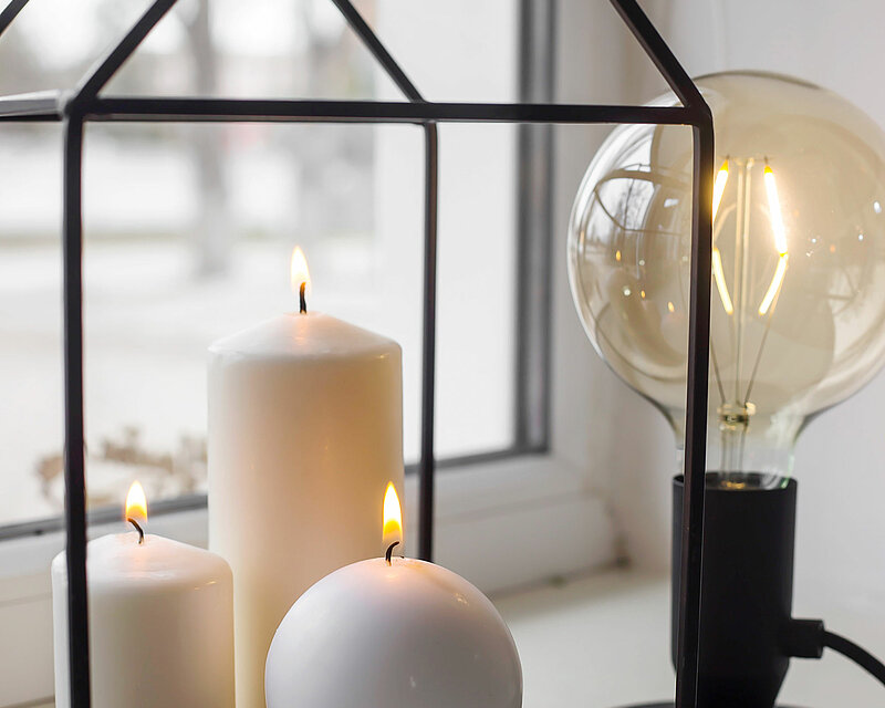 Eine weiße Kugelkerze steht zusammen mit zwei elfenbein farbenen Stumpenkerzen neben einer modernen LED Leuchte vor dem Fenster.