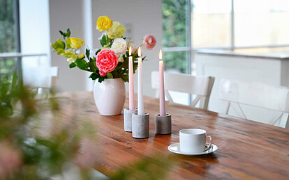 Ein gemütlicher Esstisch aus Holz mit zwei weißen Holzstühlen davor. Auf dem Tisch stehen neben einer weißen Vase mit einem Blumenstrauß und einer Kaffeetasse, noch drei Spitzkerzen in weiß und rosa in Betonkerzenhaltern.