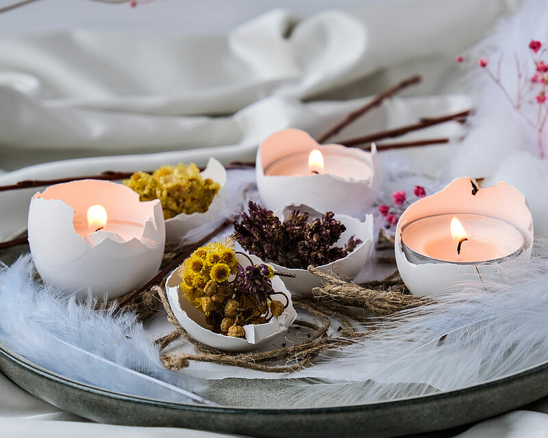 Auf einem zarten weißen Seidentuch steht ein schöner Kerzenteller, gefüllt mit weißen Federn, getrockneten Hölzern und geöffneten Eierschalen, die mit gelben Mimosenblüten dekoriert sind. In drei der halbierten Eierschalen steht jeweils ein brennendes Teelicht. Im Hintergrund erkennt man mehrere Zweige eines Baumes mit pinken Blüten. Die Szene wirkt frisch und frühlingshaft und strahlend.