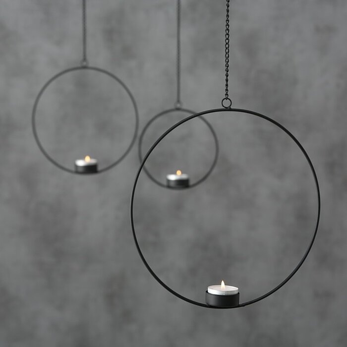 Zwei goldene Hängehülsen von Boltze mit je einem brennenden Teelicht darin, schweben in verschiedenen Höhen vor einer grauen Wand. Eine dritte Hängehülse im Hintergrund ist teilweise sichtbar und wirkt unscharf, ebenfalls mit einem brennenden Teelicht. Di