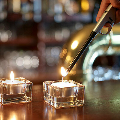 Zwei brennende Teelichter in quadratischen Teelichthaltern aus Glas auf dem Bartresen bei gemütlichem Licht. Im Hintergrund sind die Gläser und die goldene Zapfanlage der Bar verschwommen zu erkennen. Die zweite Kerze wird gerade mit einem Stabfeuerzeug angezündet.