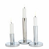 Drei brennende, weiße Stabkerzen in unterschiedlichen Höhen stehen auf glänzenden, silbernen Kerzenhaltern mit runden Sockeln. Die Kerzen strahlen einen ruhigen, schlichten und modernen Charme aus.