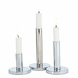Drei brennende, weiße Stabkerzen in unterschiedlichen Höhen stehen auf glänzenden, silbernen Kerzenhaltern mit runden Sockeln. Die Kerzen strahlen einen ruhigen, schlichten und modernen Charme aus.