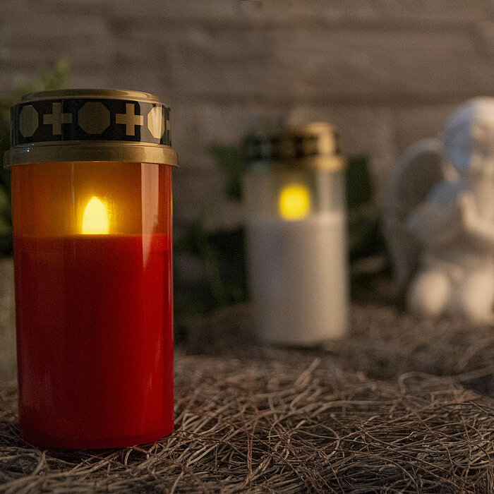 Eine rote elektronische Kerze von ARTS steht im Vordergrund auf einem natürlichen Untergrund, mit einer weiteren weißen elektronischen Kerze von ARTS im Hintergrund. Beide Kerzen haben goldene Verzierungen am oberen Rand. Im Hintergrund ist eine Engelsfig