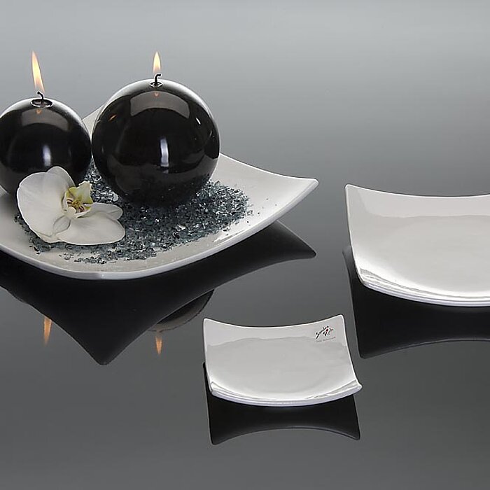 Zwei brennende schwarze Kugelkerzen stehen auf einem flachen, eckigen weißen Teller, der mit blauen Dekosteinen und einer weißen Orchideenblüte geschmückt ist. Daneben befinden sich zwei kleinere, ebenfalls eckige weiße Teller auf einer glänzenden, schwar