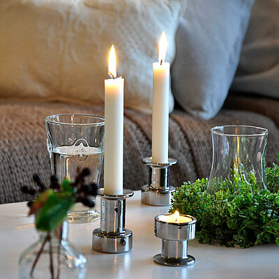 Auf einem weißen Tisch in einem gemütlichen Wohnzimmer stehen zwei silberfarbene Stabkerzenhalter mit je einer brennenden Weißen stabkerzen und ein silberner Teelichthalter mit einem brennenden Teelicht. Auf dem Tisch steht ebenfalls ein Kranz aus grünen Blättern und ein Trinkglas gefüllt mit Wasser.