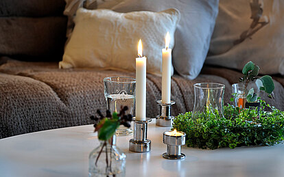 Auf einem weißen Tisch in einem gemütlichen Wohnzimmer stehen zwei silberfarbene Stabkerzenhalter mit je einer brennenden Weißen stabkerzen und ein silberner Teelichthalter mit einem brennenden Teelicht. Auf dem Tisch steht ebenfalls ein Kranz aus grünen Blättern und ein Trinkglas gefüllt mit Wasser.