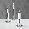 Drei weiße, brennende Stabkerzen stehen in silbernen Kerzenhaltern auf einer weißen Oberfläche. Der Hintergrund ist grau und schlicht, was die Eleganz und Schlichtheit der Szene unterstreicht. Die minimalistische Anordnung und die glänzenden Kerzenhalter 