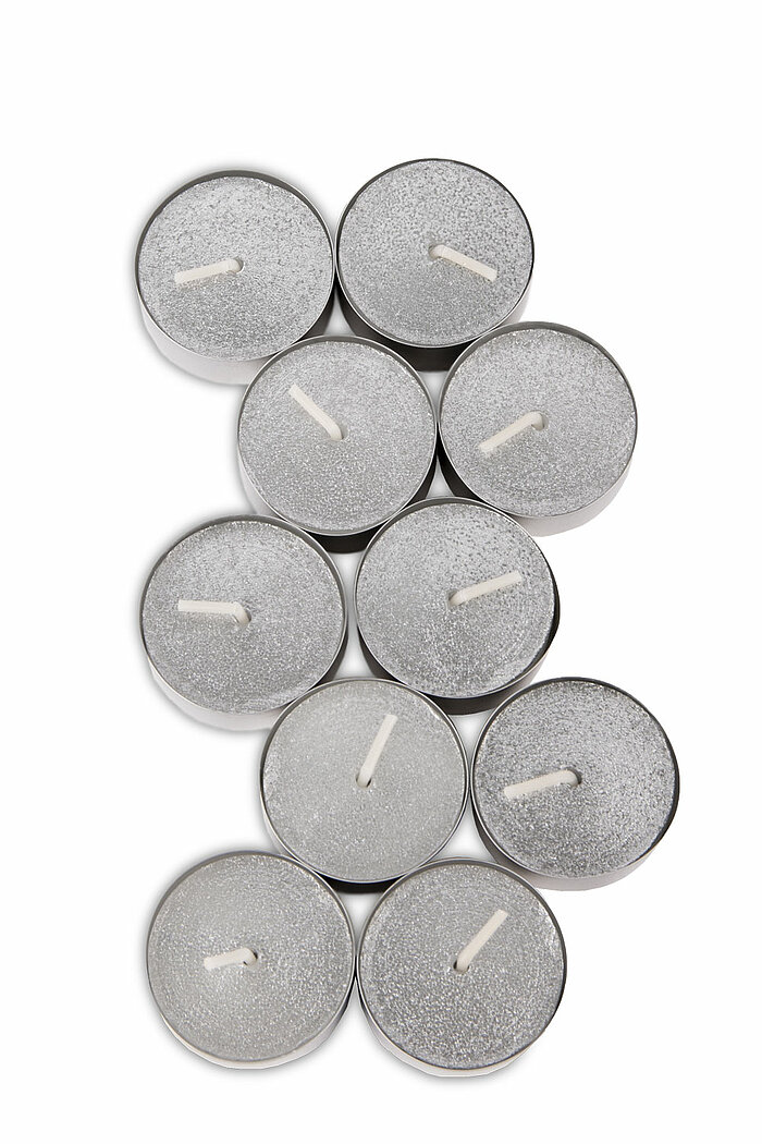 Neun silberne Teelichter, die dicht nebeneinander auf weißem Hintergrund angeordnet sind. Die Teelichter haben eine glänzende, leicht glitzernde Oberfläche und befinden sich in einem metallischen Gehäuse. Sie sind alle unangezündet und zeigen ihre Dochte 