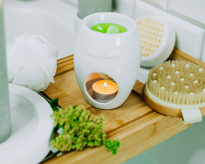 Auf einer zum teil gefüllten Badewanne steht auf den Rändern ein Holztablett. Auf dem Holztablett befindet sich eine weisse ovale Duftlaterne von Bolsius mit einem brennenden Teelicht und gefüllt mit Wachsmelts. Neben der Duftlampe befinden sich einige Wellness Sachen und dahinter eine grüne brennende Rustikale Stumpenkerze.