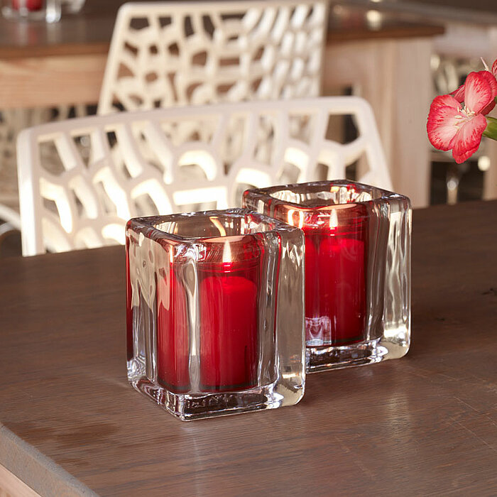 Ein kleiner Tisch in einem stilvollen Restaurant ist mit einem Bolsius-Teelicht in einem roten Glas dekoriert. Daneben stehen eine kleine Edelstahlkanne und eine weiße Vase mit orangefarbenen Blumen. Der Tisch ist mit einer hellen Tischdecke, Holzbrettern