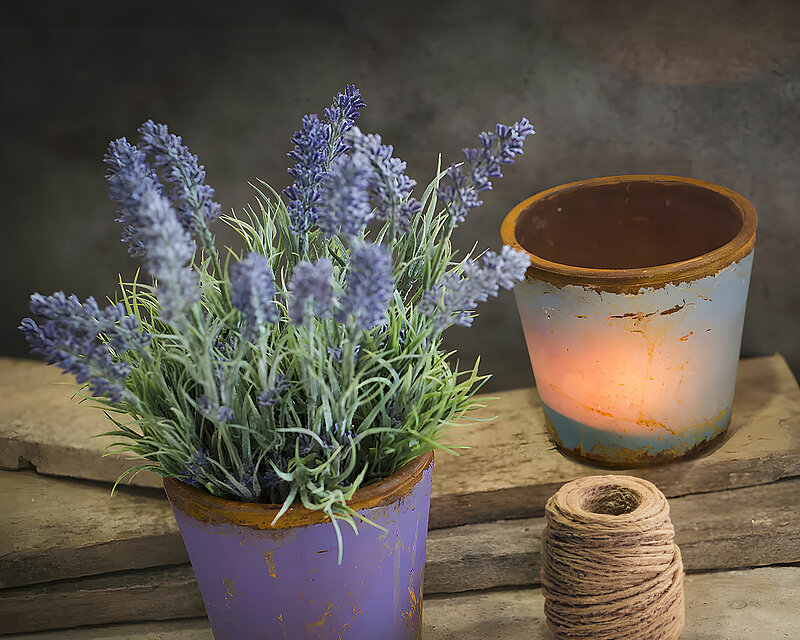 Auf einer rustikalen Holzplatte steht eine Glasvase, der "Vintage Flower Pot" in der Farbe Lavendel von Sandra Rich. Daneben liegt eine braune Baumwollkordel, fein säuberlich aufgerollt. In der Vase befinden sich einige Zweige mit saftigem Lavendel. Auf einem Holzbrett hinter der Vase steht ebenfalls eine Glasvase, der "Vintage Flower Pot" in der Farbe Eisblau. Das gesamte Bild strahlt Ruhe und Rustikalität aus und ist in sanftes Licht getaucht.