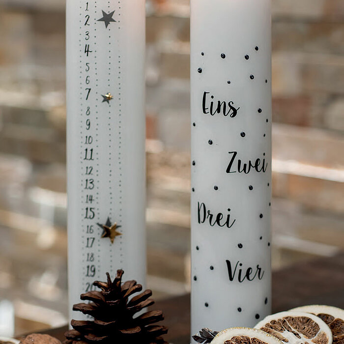 Zwei brennende Stabkerzen des Herstellers Wenzel, die auf einem Tisch dekoriert sind. Eine Kerze ist mit einem Sternenmuster und Zahlen versehen, die als Countdown bis Weihnachten zählen. Die andere Kerze ist mit den Zahlen Eins, Zwei, Drei und Vier sowie