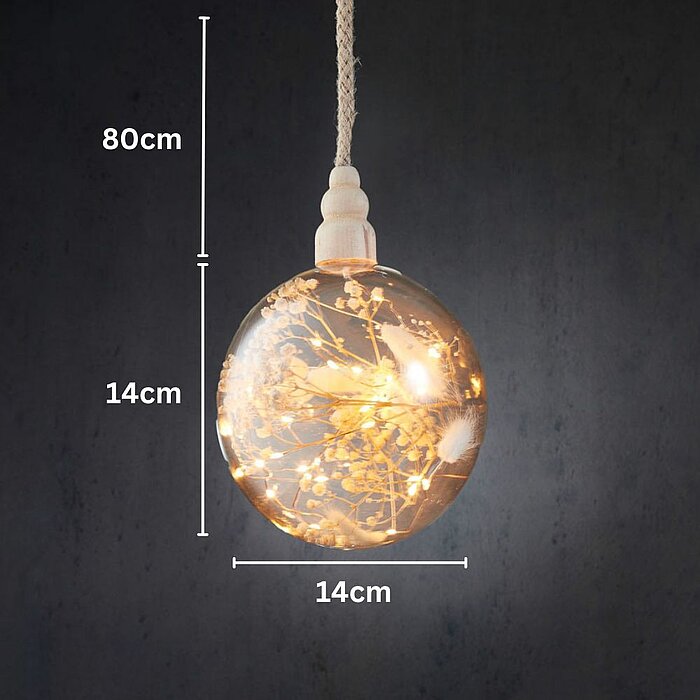 Eine kugelförmige Hängelampe leuchtet in warmem Licht. In ihrem Inneren sind zarte, getrocknete Blumen arrangiert, die von einem feinen Netz aus kleinen Glühbirnen umgeben sind. Die Lampe hängt vor einem dunkelgrauen Hintergrund, was den filigranen Charak