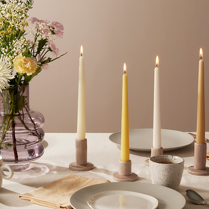 Vier brennende Tafelkerzen von Bolsius in Weiß und Gelb stehen in schlichten, zylinderförmigen Kerzenhaltern auf einem gedeckten Esstisch. Die Kerzen sind neben einem violetten Glasgefäß mit verschiedenen Blumen arrangiert. Der Tisch ist mit weißem Geschi
