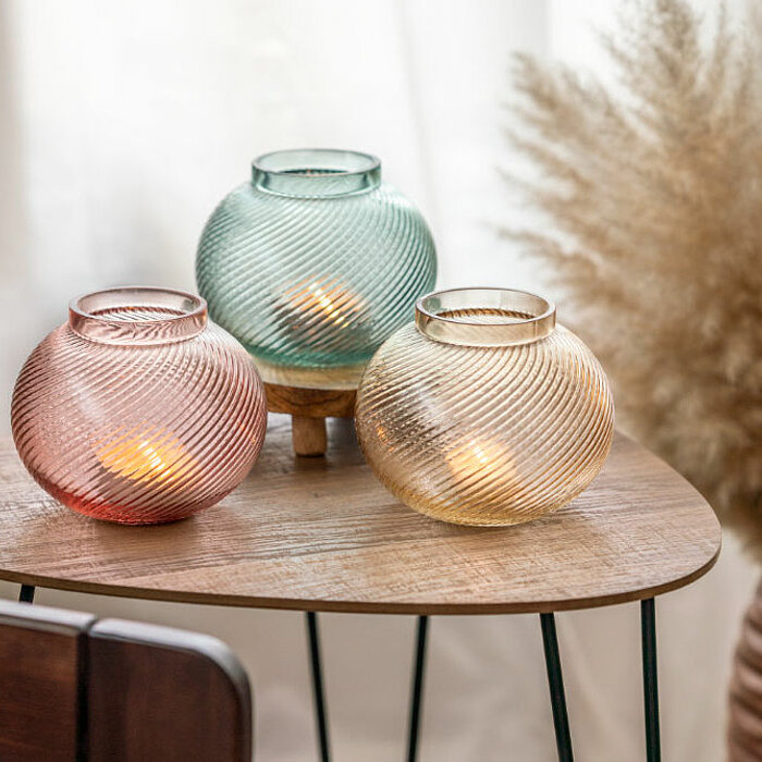 Drei Sandra Rich Windlichter in sanften Farben stehen auf einem runden Holztisch: eines in Rosa, eines in Grün und eines in Hellgelb. Die Lichter verbreiten ein warmes, gemütliches Licht. Im Hintergrund sind getrocknete Pflanzenarrangements in einer Vase 