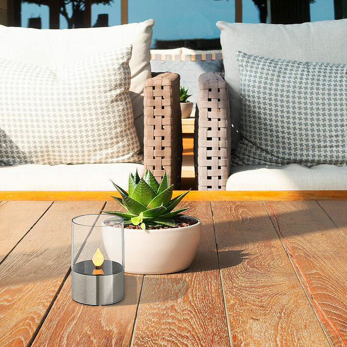 Auf einem hölzernen Terrassentisch steht eine brennende Kerze in einem transparenten Glasbehälter. Neben der Kerze befindet sich ein Pflanztopf mit einer kleinen grünen Sukkulente. Im Hintergrund sind zwei bequeme Rattansessel mit gemusterten Kissen zu se