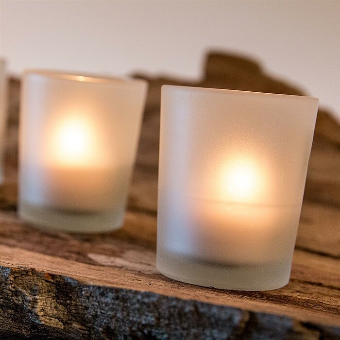 Zwei brennende Teelichter in Satin-Glasbehältern vom Hersteller "Sandra Rich" stehen nebeneinander auf einer glänzenden, roten Oberfläche. Im Hintergrund sind kunstvoll geformte Drahtkugeln und eine hölzerne Wand zu sehen, was eine warme und gemütliche At