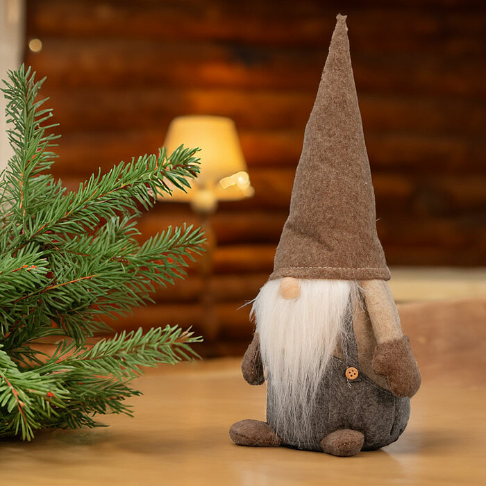 Zwei Stoff-Wichtel des Herstellers "Edelman", mit langen weißen Bärten und spitzen Hüten in Brauntönen, stehen nebeneinander auf einem schwarzen Regal. Im Hintergrund sind verschwommen ein modernes Sofa und dekorative Elemente zu sehen, was eine gemütlich