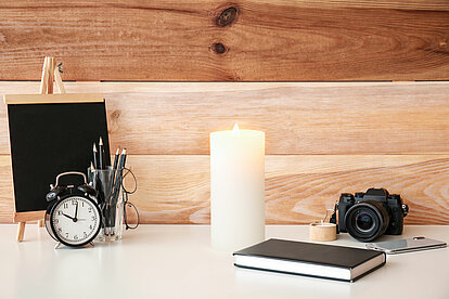 Auf dem Bild ist ein weisser Tisch zu sehen mit einer weissen brennenden Stumpenkerze. Daneben liegt eine schöne Fotokamera und auf dem Tisch steht ein schwarzer Wecker sowie eine kleine Kreidetafel.