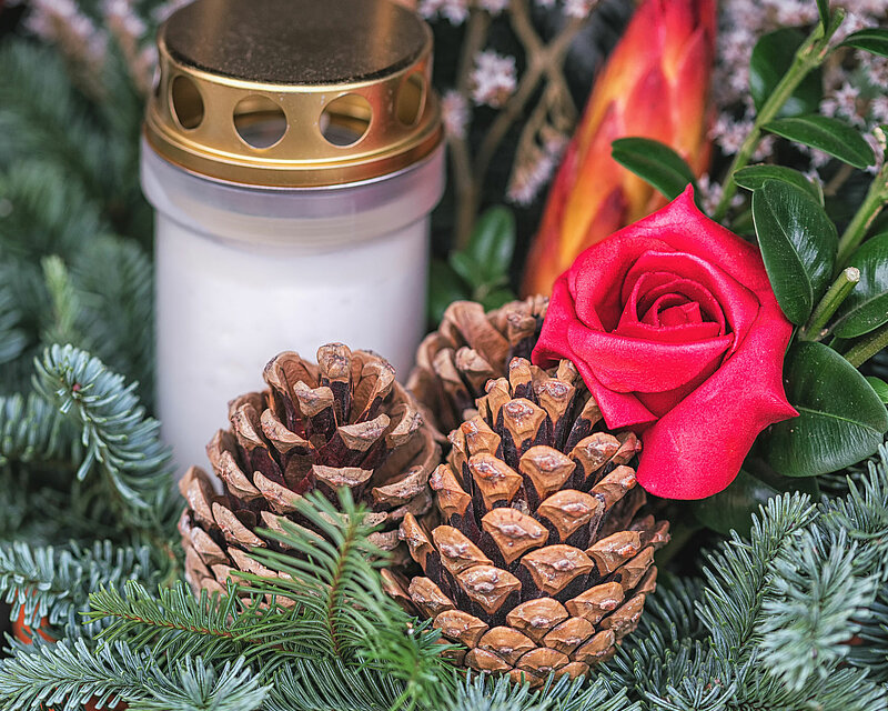In einer Tonschale stecken mehrere grüne Tannenzweige und Tannenzapfen, die eine natürliche, rustikale Atmosphäre schaffen. Dazwischen befinden sich eine rote Rose und ein weißes Grablicht mit goldenem Deckel, das einen eleganten Akzent setzt. Im Hintergrund sind einige weiße Blüten zu erkennen, die vom natürlichen, hellen Tageslicht angestrahlt werden und dem Arrangement eine friedliche Stimmung verleihen.