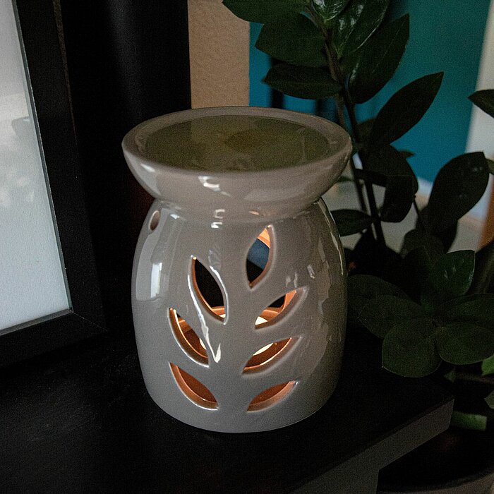 Ein cremefarbener Keramik-Duftölbrenner mit ausgeschnittenen Blattformen steht auf einem dunklen Holzregal. Im Inneren des Brenners brennt ein Teelicht, das durch die Aussparungen ein warmes Licht erzeugt. Im Hintergrund sind grüne Pflanzen und unscharfe 