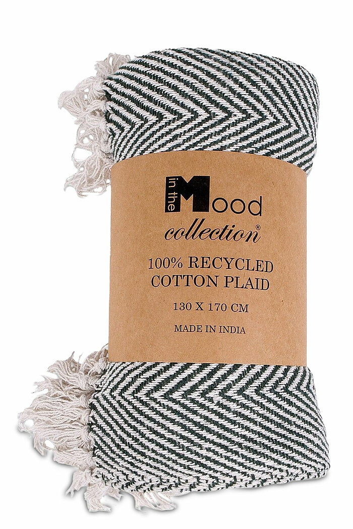 Eine zusammengerollte Decke aus der "Mood Collection". Sie besteht zu 100% aus recycelter Baumwolle und hat die Maße 130 x 170 cm. Das Muster der Decke zeigt ein schwarzes und weißes Fischgrätenmuster. Die Enden der Decke sind mit weißen Fransen versehen.