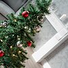 Ein elegant dekorierter Weihnachtsbaum mit grünen Zweigen, roten und silbernen Kugeln sowie warmen Lichterketten steht im Bildvordergrund. Im Hintergrund sieht man einen weißen Kaminsims, auf dem eine brennende weiße Kerze und silberne Dekorationen stehen