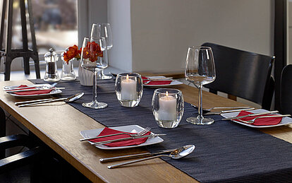 bolsius-kerzen-gastronomie-impression-2023-1 Die Kerzen Gastronomie setzt auf eine stilvolle Tischdekoration in einem Restaurant, die mit weißen Tellern und roten Servietten anspruchsvoll gestaltet ist. Auf dem Tisch steht eine weiße Vase mit roten Blumen und leere Weingläser, begleitet von zwei brennenden Relight Kerzen in einem ovalen Glashalter, die eine gemütliche und einladende Stimmung verbreiten.
